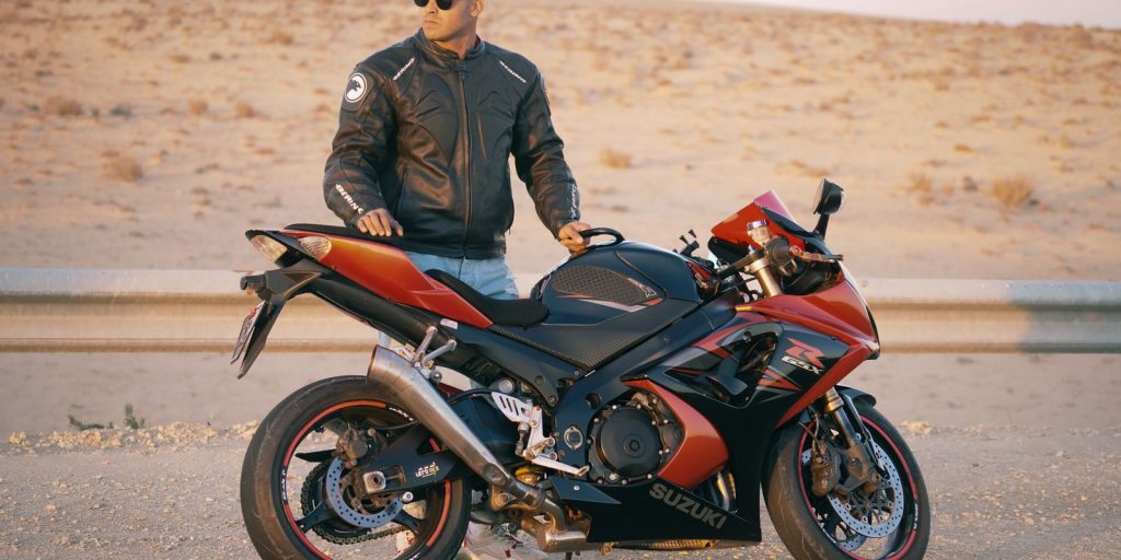 Hombre con chaqueta de cuero y gorra posando junto a una moto deportiva en una carretera desértica
