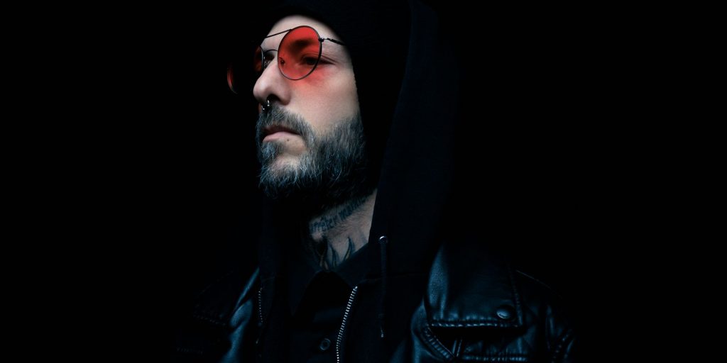 Retrato melancólico de un hombre con chaqueta de cuero negra y gafas de tono rojo sobre fondo oscuro