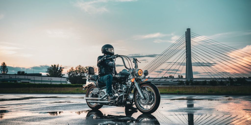 Motociclista con equipo de cuero en una carretera mojada con un puente al fondo al atardecer