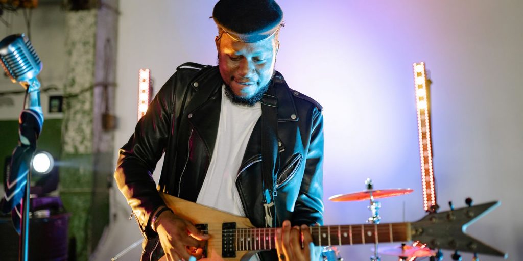 Guitarrista tocando una guitarra eléctrica en el escenario con luces vibrantes, vistiendo chaqueta de cuero y gorra plana